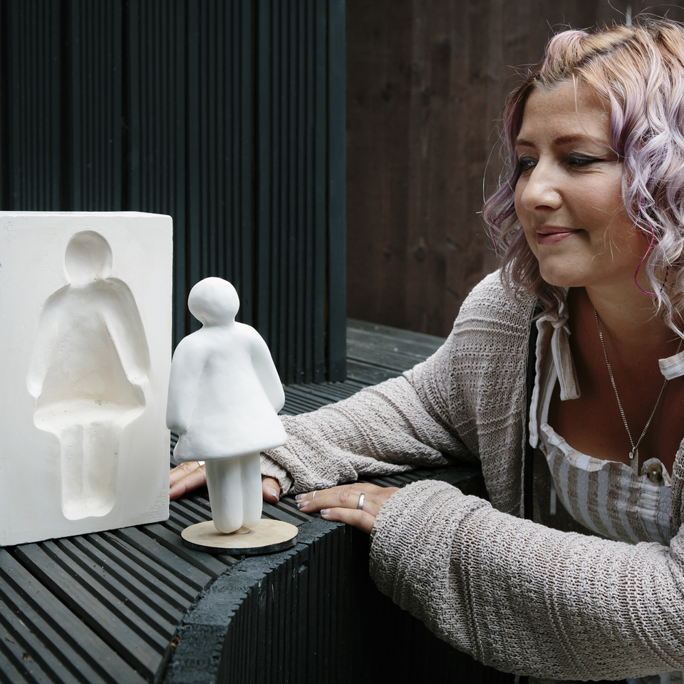 A woman with wavy, pastel-colored hair smiles while looking at a small white sculpture of a person and its matching mold on a dark, slatted surface.